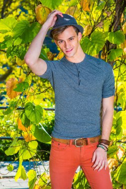 Genç adamın portresi. Gri uzun kollu, yuvarlak taçlı Henley gömlekli, kırmızı kotlu, yeşil bitkilerin yanında duran, yün fedora şapkalı, gülümseyen, sana bakan genç bir adam.