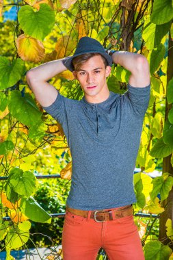 Gri uzun kollu, yuvarlak taçlı Henley gömlekli, kırmızı kotlu, yün bir Fedora şapkalı genç yakışıklı bir adam yeşil çiçeklerin yanında duruyor, kafasının arkasından ellerini kaldırıyor ve sana bakıyor..