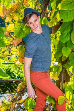 Sonbahar Öğleden Sonra Eğlenen Adam. Gri uzun kollu, yuvarlak taçlı Henley gömlekli, kırmızı kotlu, yün bir Fedora şapkalı genç yakışıklı bir adam yeşil bitkilerin yanında duruyor ve sana bakıyor.