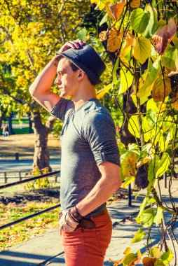 Seni Bekleyen Adam. Yuvarlak gömlekli, kırmızı kot pantolonlu, yünlü Fedora şapkalı gri uzun kollu genç bir adam altın sonbahar yapraklarının yanında sabırla ileriye bakıyor..