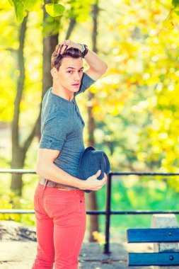 Bir adam seni arıyor. Uzun kollu yuvarlak bir gömlek, kırmızı kot pantolon, yünlü bir Fedora şapkası tutan, saçlarına dokunan genç yakışıklı bir adam, altın sonbahar yapraklarının yanında duruyor, geriye bakıyor.