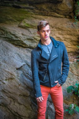 Mavi dar bir ceket, kırmızı kot pantolon giymiş genç bir adam kayaların yanında duruyor, dinleniyor ve düşünüyor.