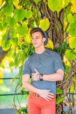 Genç adam. Gri uzun kollu, yuvarlak bir Henley gömlek, kırmızı kot, yün bir Fedora şapkası giyen, genç yakışıklı bir adam büyük bir yaprağın yanında duruyor, elleri karnında, kaşlarını çatmış, düşünüyor..