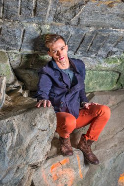 Vücudunun üzerine eğilmiş, genç bir adam kayalardan bir mağarada çömeliyor, üzgün..