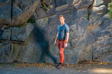 Gri, uzun kollu, yuvarlak bir Henley gömlek, kırmızı kot pantolon, kahverengi deri ayakkabılar giyen genç bir adam büyük kayaların yanında duruyor, gülümsüyor ve sana bakıyor.