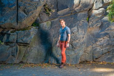 Gri, uzun kollu, yuvarlak bir Henley gömlek, kırmızı kot pantolon, kahverengi deri ayakkabılar giyen genç bir adam büyük kayaların yanında duruyor, gülümsüyor ve sana bakıyor.
