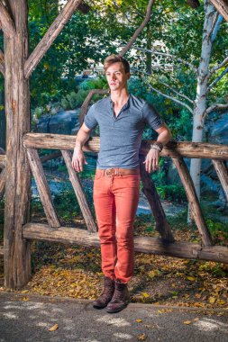 Uzun kollu, yuvarlak taçlı Henley gömlek, kırmızı kot pantolon, kahverengi çizme ayakkabı giyen genç bir adam, tahta bir çite karşı dimdik duruyor, sıkı düşünüyor.