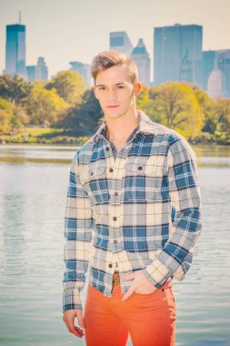 Genç adamın portresi. Uzun kollu, desenli gömlekli, kırmızı pantolonlu, bir eli cebinde genç bir adam gölün kenarında duruyor. Arka plandaki yüksek iş binaları. Filtre etkisi.