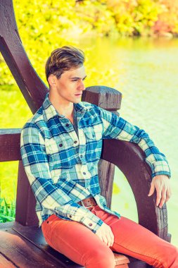 Genç adam dinleniyor. Uzun kollu, desenli gömlekli, kırmızı pantolonlu genç bir adam su kenarında seni bekliyor. filtrelenmiş etki