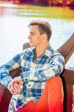 Genç Yakışıklı Adam 'ın portresi. Uzun kollu, desenli gömlekli, kırmızı pantolonlu genç bir adam nedense su kenarında bankta oturuyor ve kendinden emin bir şekilde ileriye bakıyor. filtrelenmiş etki