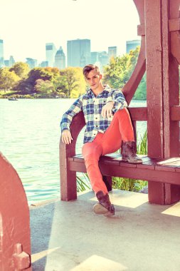 Uzun kollu, desenli gömlekli, kırmızı pantolonlu, kahverengi çizmeli genç bir adam nedense gölün kenarında oturmuş düşünüyor. Arka plandaki şehir binaları. Retro filtrelenmiş efekt. / Par 'daki genç adam