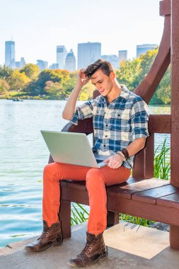 Genç öğrenci dışarıda okuyor. Uzun kollu, desenli bir gömlek, kırmızı pantolon, kahverengi botlar, başını kaşıyan bir el, genç bir adam öğleden sonra göl kenarında dizüstü bilgisayarda çalışıyor. Arka planda yüksek iş binaları.