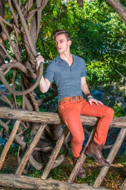 Gri uzun kollu, yuvarlak taçlı Henley gömlekli, kırmızı kot pantolonlu, kahverengi deri ayakkabılı, genç bir adam tahta bir çitin üzerinde oturuyor, elinde kavisli bir rattan, seni bekliyor..