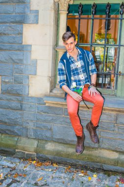 Mavi ve beyaz desenli gömlek, mavi kapüşonlu yelek, kırmızı kot pantolon ve kahverengi deri ayakkabı giymiş genç bir adam pencerenin kenarında oturuyor, elinde beyaz bir çiçek, seni bekliyor..