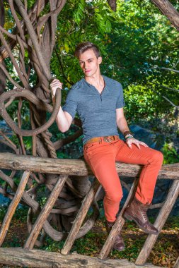 Gri uzun kollu, yuvarlak taçlı Henley gömlek, kırmızı kot pantolon, kahverengi deri ayakkabı giymiş, genç bir adam ahşap bir çitin üzerinde oturuyor, elinde kavisli bir rattan, sana bakıyor..