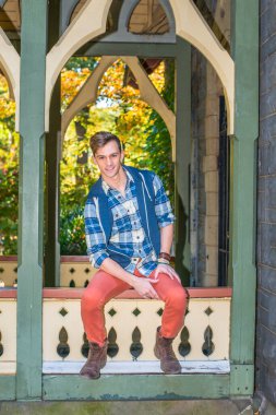 Genç adam dışarıda dinleniyor. Mavi ve beyaz desenli gömlek, mavi kapüşonlu yelek, kırmızı kot pantolon ve kahverengi deri ayakkabılar giyen genç bir adam bir köşkte seni bekliyor..