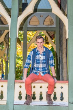 Genç adam dışarıda dinleniyor. Mavi ve beyaz desenli gömlek, mavi kapüşonlu yelek, kırmızı kot pantolon ve kahverengi deri ayakkabılar giyen genç bir adam bir köşkte seni bekliyor.