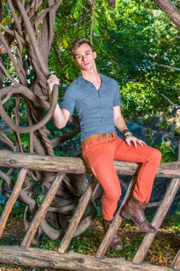 Gri uzun kollu, yuvarlak taçlı Henley gömlek, kırmızı kot pantolon, kahverengi deri ayakkabı giymiş, genç bir adam ahşap bir çitte oturuyor, elinde kavisli bir rattan, dışarıda dinleniyor..