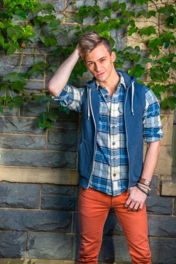 Adam dışarıda dinleniyor. Mavi ve beyaz desenli gömlek, mavi kapüşonlu yelek, kırmızı kot pantolon, bir eli kaşınan kafa, genç adam yeşil sarmaşık yapraklı bir duvarın yanında duruyor, dinleniyor ve düşünüyor.