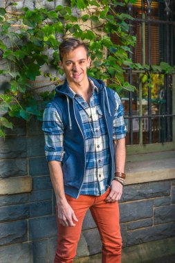 Adam dışarıda dinleniyor. Mavi ve beyaz desenli gömlek, mavi kapüşonlu yelek, kırmızı kot pantolon giyen genç bir adam pencerenin yanında duruyor ve yeşil sarmaşık yapraklı bir duvar.