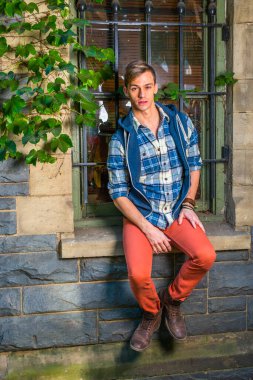 Genç adam dışarıda dinleniyor. Mavi ve beyaz desenli gömlek, mavi kapüşonlu yelek, kırmızı kot pantolon ve kahverengi deri ayakkabı giyen genç bir adam pencerenin kenarında seni bekliyor.