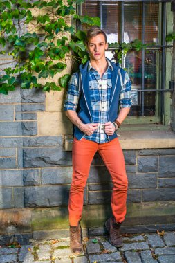 Sıradan erkek modası. Mavi ve beyaz desenli gömlek, mavi kapüşonlu yelek, kırmızı kot pantolon, kahverengi deri ayakkabı giyen genç bir adam, pencerenin köşesinde duruyor ve yeşil sarmaşık yapraklı bir duvar.