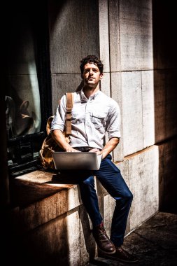 Giyen Beyaz tişört, Mavi pantolon, kahverengi ayakkabı, pencere çerçevesi dizüstü bilgisayar üzerinde çalışan güneş altında kampüste oturan sarı omuz çantası taşıyan genç Amerikalı adam, New York'ta eğitim düşünme