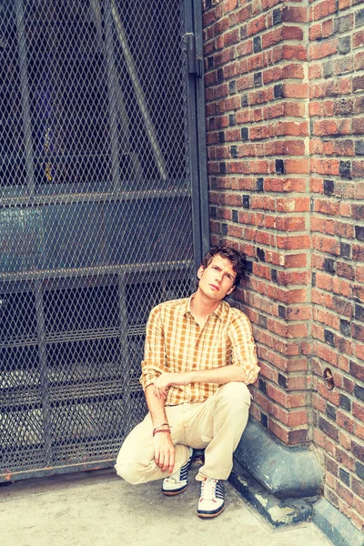 Concept of lonely man thinking about love. Wearing yellow patterned shirt, pants, sneakers, a young European guy squatting at corner on street in New York, looking up, sad, thinking. I missing You