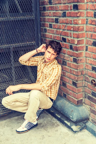 Concept of lonely man thinking about love. Wearing yellow patterned shirt, pants, sneakers, a young European guy squatting at corner on street in New York, sad, thinking after busy working day