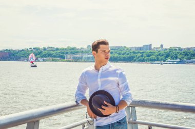 Beyaz gömlekli genç adam, elinde Fedora şapkası, güneş gözlüğü New Jersey 'in karşısındaki Hudson Nehri' nin yanında duruyor, geriye dönüp ileriye bakıyor. Arka planda küçük bir tekne. Boşluğu Kopyala