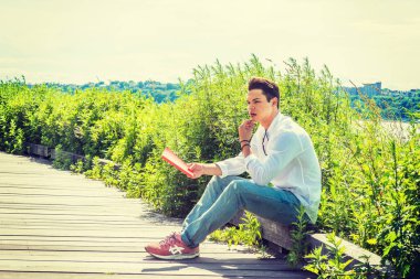 Park 'ta okuyorum. Beyaz gömlek, kot pantolon, spor ayakkabı, yakasında güneş gözlüğü, New York 'ta Hudson Nehri' nin kenarında oturan genç bir adam, kırmızı kitap tutan bir el, çeneye dokunan, okuyan, düşünen