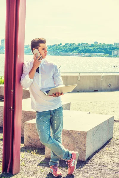 College Student Studying in New York. Wearing white shirt, jeans, sneakers, a young guy standing against pole by Hudson River, working on laptop computer, talking on phone.