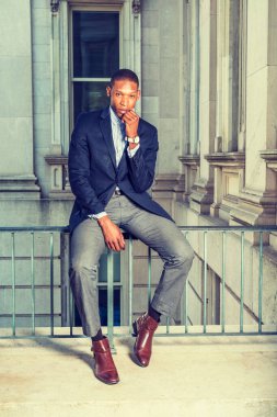 Siyah ceket, gri pantolon, kahverengi deri ayakkabı, kol saati, Afrika kökenli Amerikalı iş adamı klasik ofis binasında korkuluklarda oturuyor, çeneye yumruk atıyor, düşünüyor..