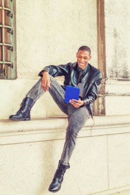 Modern okuma. Siyah deri ceket, çizgili atlet, kot pantolon, botlar, el bilgisayarı, New York 'ta duvara yaslanmış, gülümseyen, okuyan bir Afrikalı Amerikalı.
