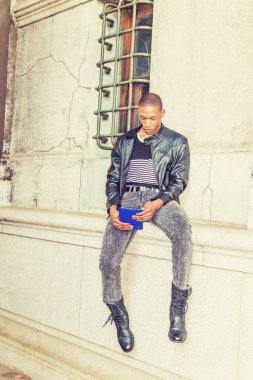 Modern okuma. Siyah deri ceket, çizgili atlet, kot pantolon, çizme, el bilgisayarı, New York 'ta duvara yaslanmış kitap okuyan genç bir zenci.