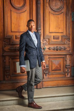 Siyah ceket, gri pantolon, kahverengi deri ayakkabı, dizüstü bilgisayar taşıyan genç bir Afro-Amerikan işadamı klasik ofis kapısından aşağı iniyor, ileriye bakıyor..