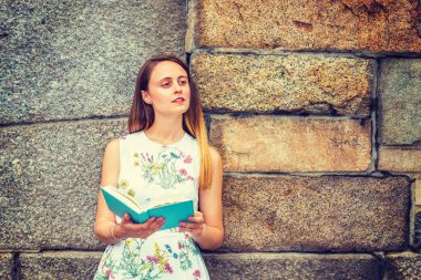 Amerikan üniversite öğrencisinin portresi. Çiçek desenli, kolsuz, beyaz bir elbise giyen, elinde yeşil bir kitap tutan, kayalık duvarın yanında duran, gözlerini kısarak okuyan, düşünen güzel bir kız.