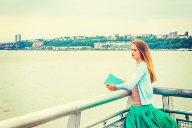Unutulmaz okuma. Açık mavi hırka, yeşil etek, kitap, New Jersey 'in karşısındaki Hudson Nehri' nin yanında metal çitin yanında duran Amerikalı bir kadın..