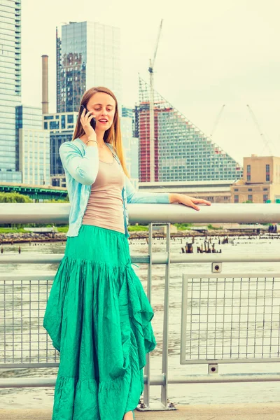 私たちの現代生活の技術 ニューヨークの桟橋にある金属製の柵のそばに立っている美しいアメリカ人女性携帯電話でリラックスして話しています — ストック写真