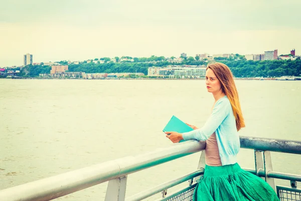 Unutulmaz okuma. Açık mavi hırka, yeşil etek, kitap, New Jersey 'in karşısındaki Hudson Nehri' nin yanında metal çitin yanında duran Amerikalı bir kadın..