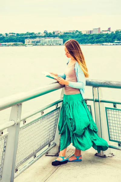 Açık mavi hırka, yeşil etek, sandalet giyen Amerikalı bir üniversite öğrencisi New Jersey 'de Hudson Nehri kıyısında metal çitlerin yanında durmuş kitap okuyor..