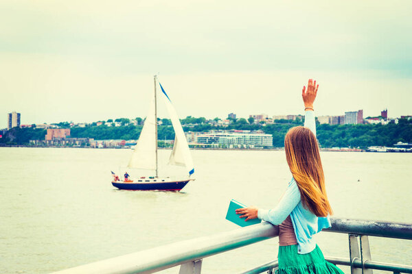 Welcome You. A girl with long blonde hair, wearing cardigan, skirt, holding book, standing by Hudson River in New York, looking at boat in water., raising arm, waving hand. Back view