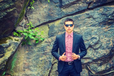 Siyah takım elbise, kırmızı, beyaz desenli fanila, güneş gözlüğü, kol saati, kısa saç kesimi, kayalık duvarın yanında duran genç seksi bir adam, gizemli bir şekilde sana bakıyor..