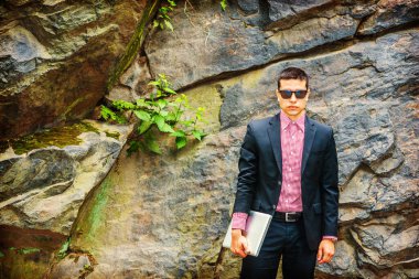 Siyah takım elbise, kırmızı, beyaz desenli fanila, güneş gözlüğü, kısa saç kesimi, dizüstü bilgisayar taşıyan, kayaların yanında duran, gizemli bir şekilde sana bakan bir öğrenci.