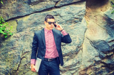 Siyah takım elbise, kırmızı, beyaz desenli fanila, güneş gözlüğü, kısa saç kesimi, dizüstü bilgisayar, dışarıda duran genç bir profesyonel, cep telefonuyla konuşuyor..