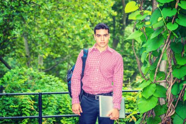 Okula geri dön. Şablon gömlekli, omuz çantalı, dizüstü bilgisayarlı, kampüste ormanda dikilen bir üniversite öğrencisi, kendinden emin bir şekilde ileriye bakıyor. Sis, arka planda çiseleme.