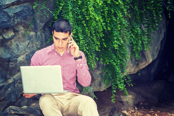 一位身穿红白相间图案衬衫的年轻大学生 靠着绿叶的岩石坐在校园里看书 在笔记本电脑上工作 一边打电话 — 图库照片