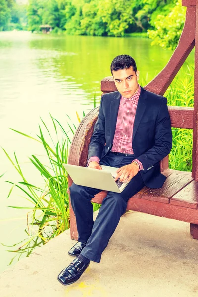 在和平环境中努力工作 穿着黑色西服 穿着红白相间的衬衫 一个年轻的大学生坐在校园湖边 在笔记本电脑上工作 — 图库照片