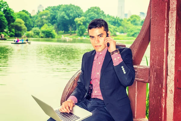 在和平的环境中工作 年轻的大学生坐在湖边 在笔记本电脑上工作 同时在手机上聊天 背景上的小船 现代假期 — 图库照片