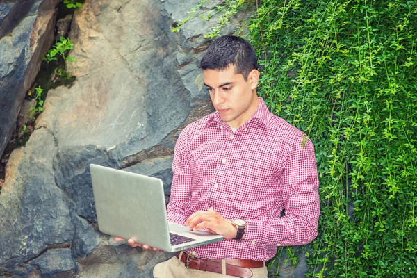 在外面念书一位年轻的大学生身穿红色图案的长袖衬衫 戴着腕表 靠着绿叶的岩石站着 低头看 在笔记本电脑上工作 — 图库照片